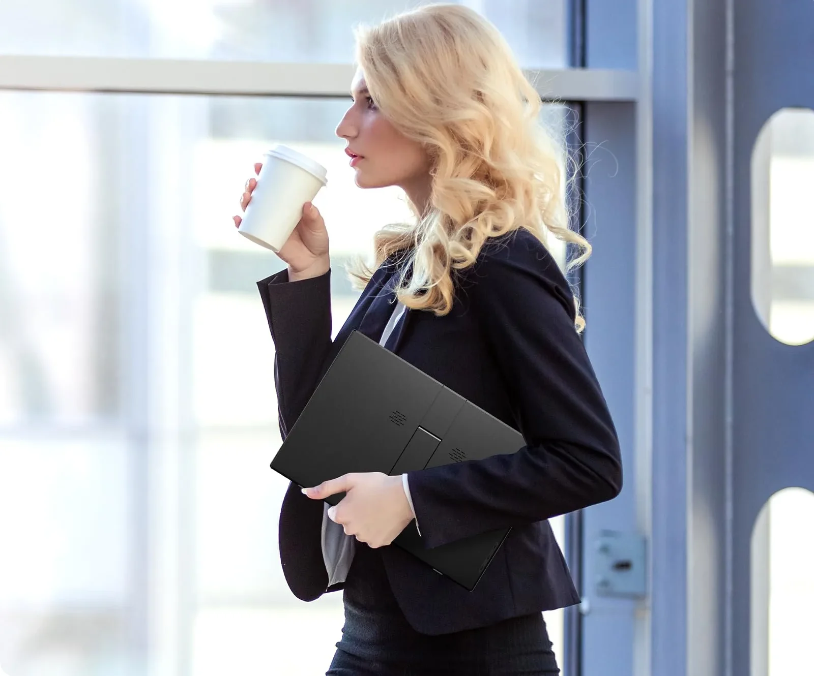 A woman in a dark blazer carrying a slim YXK portable monitor under her arm while holding a coffee